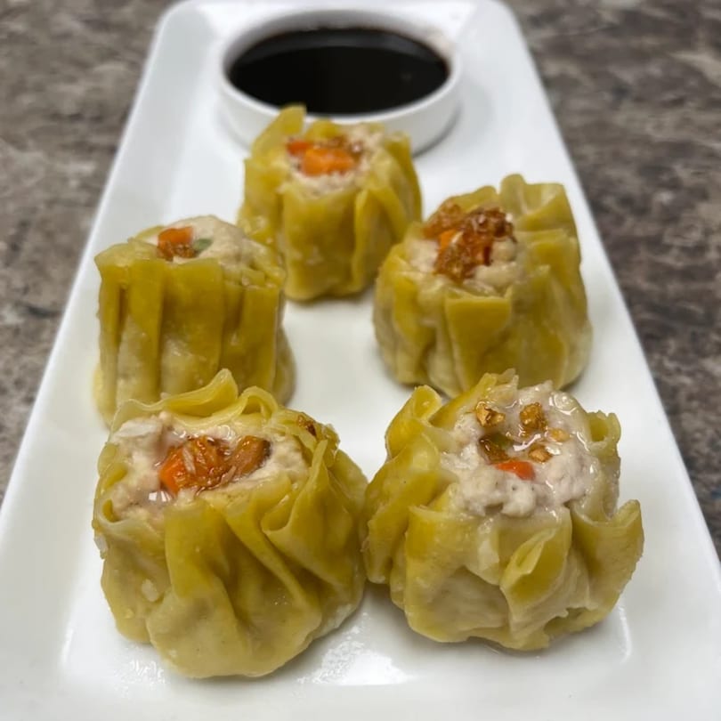 Five steamed Thai dumplings, topped with carrots and spices, served with a side of dark sauce.