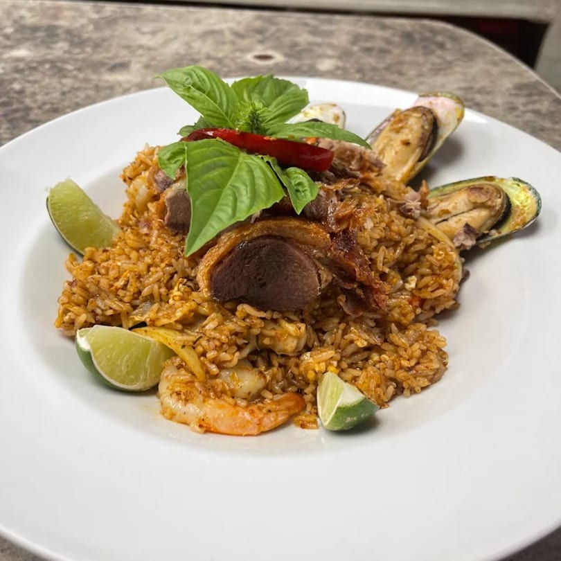 Thai fried rice with duck, shrimp, mussels, basil, red chili, and lime wedges on a white plate.